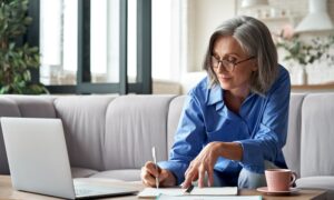 Stylish mature older woman working from home on laptop taking notes.