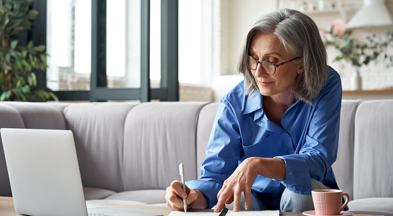 Stylish mature older woman working from home on laptop taking notes.